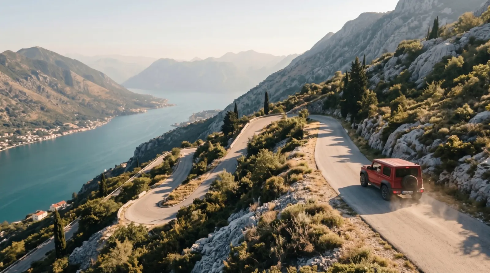 A red SUV climbs mountain hairpins above a turquoise bay in Montenegro