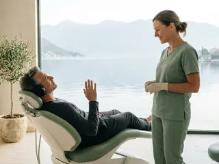 A patient wearing headphones raises his hand as the sign for a pause; the dentist stops and smiles.